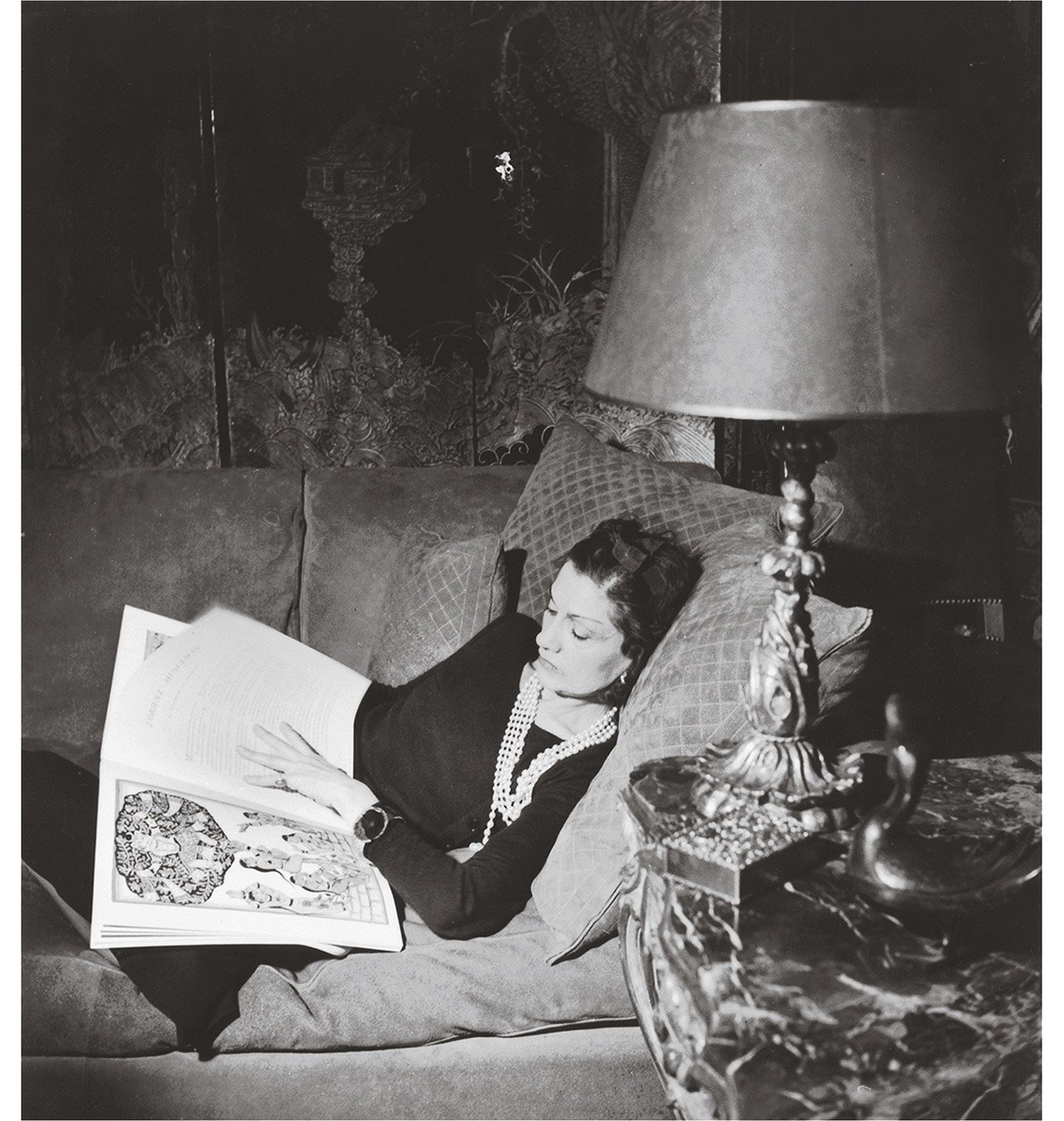 Portrait of designer Chanel seated on a couch with a book, wearing a multi-strand pearl necklace