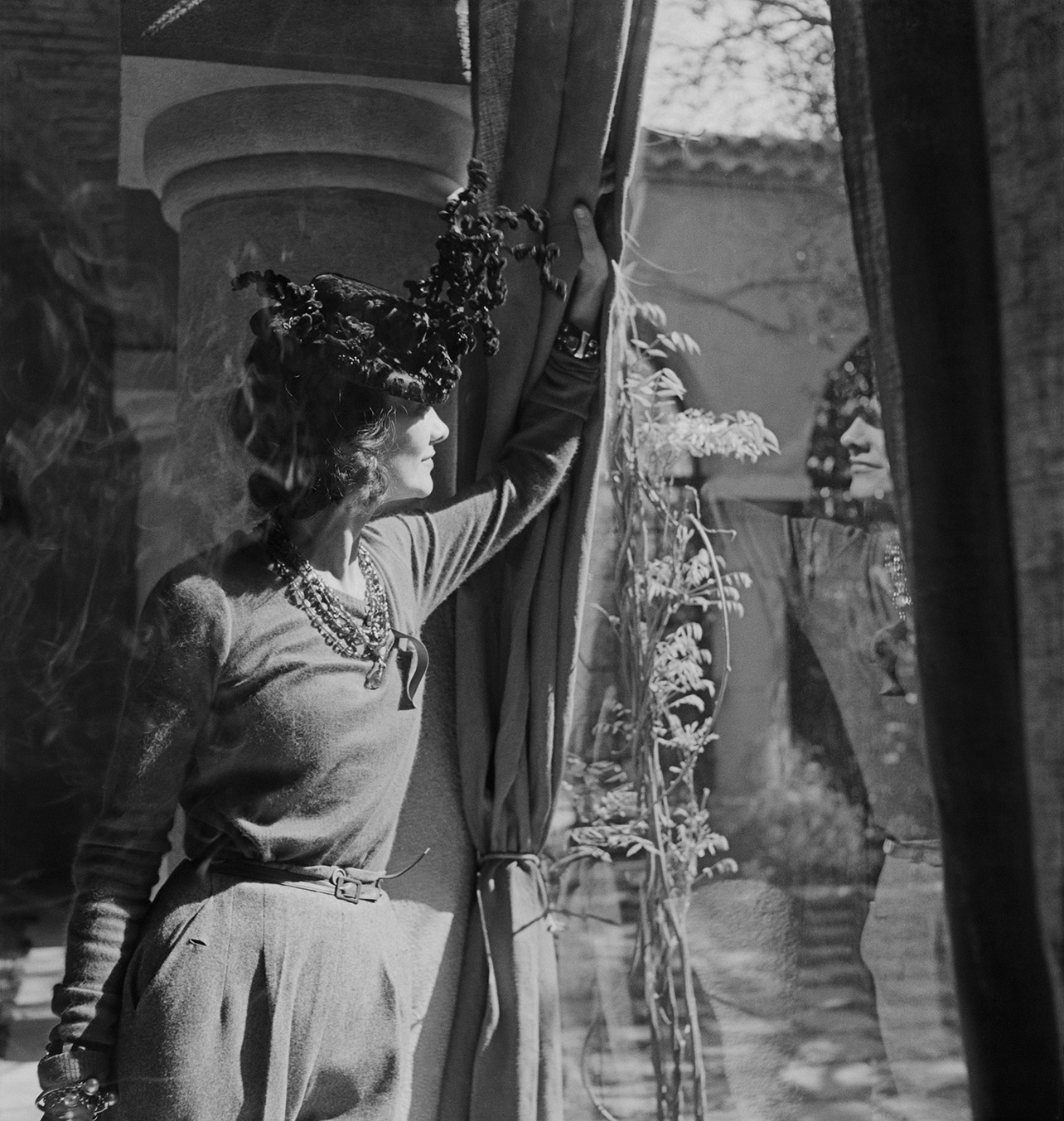 Portrait of designer Chanel posing by a large sunny window, gazing out at the garden view