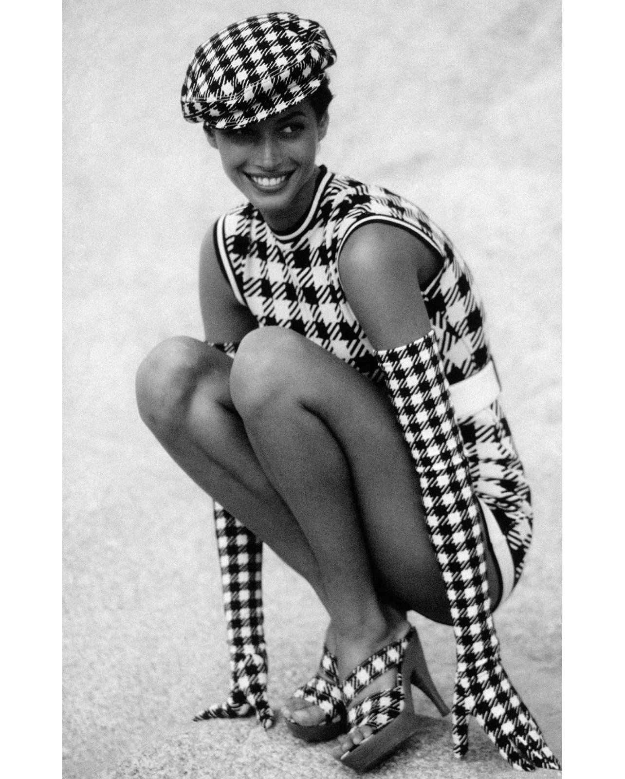 Black and white image of a fashion model posing in a coordinated patterned outfit with matching hat, dress, gloves, and heels