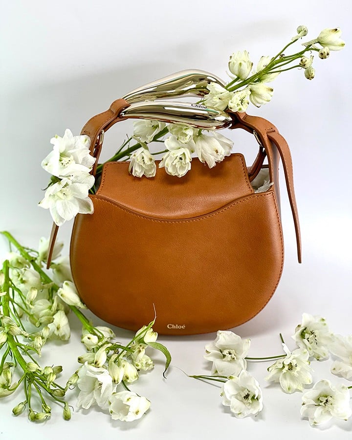 Chloé’s Kiss Leather Hobo Bag in Arizona brown with a semicircular silhouette, abstract bar handle, and top magnetic flap closure.