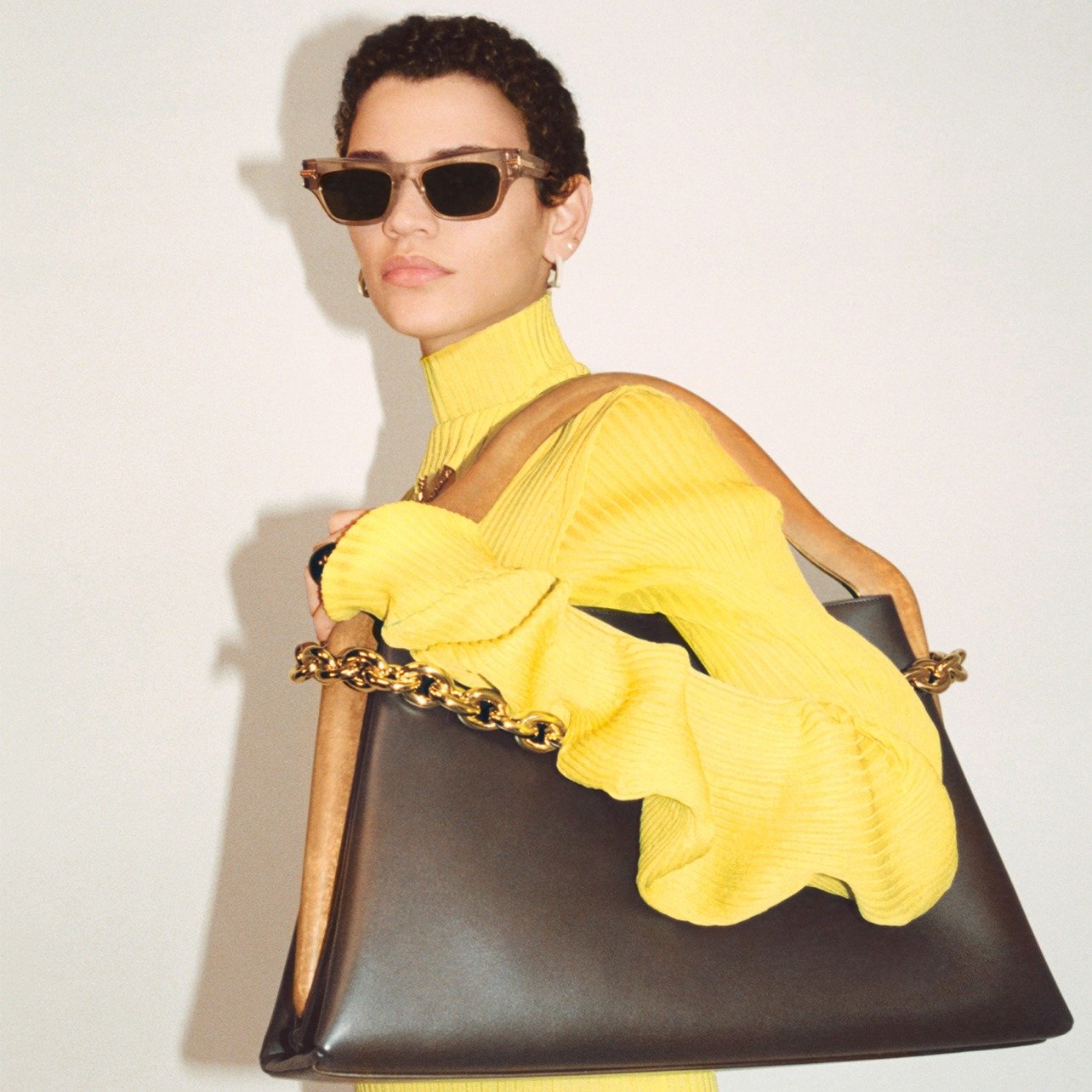 Woman poses in a high-neck yellow sweater dress while holding a large shoulder bag with gold chain detailing