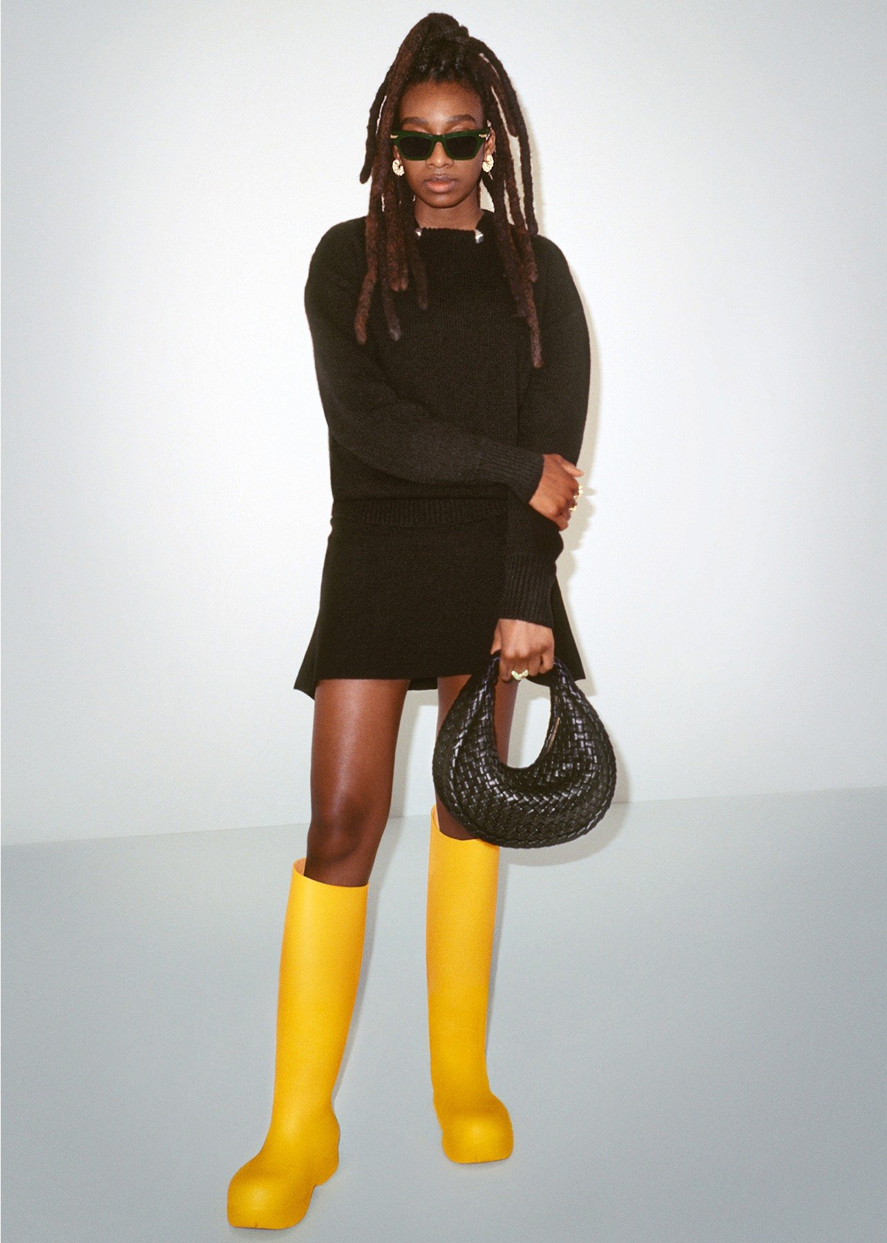 Model poses wearing a black sweater, black skirt, yellow knee-high rubber boots and holding a black woven top handle bag