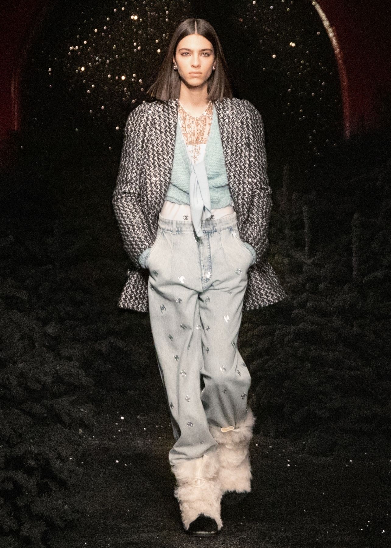 Woman pictured on runway wearing a tweed jacket, light blue blouse, light blue trousers and white fur ankle boots