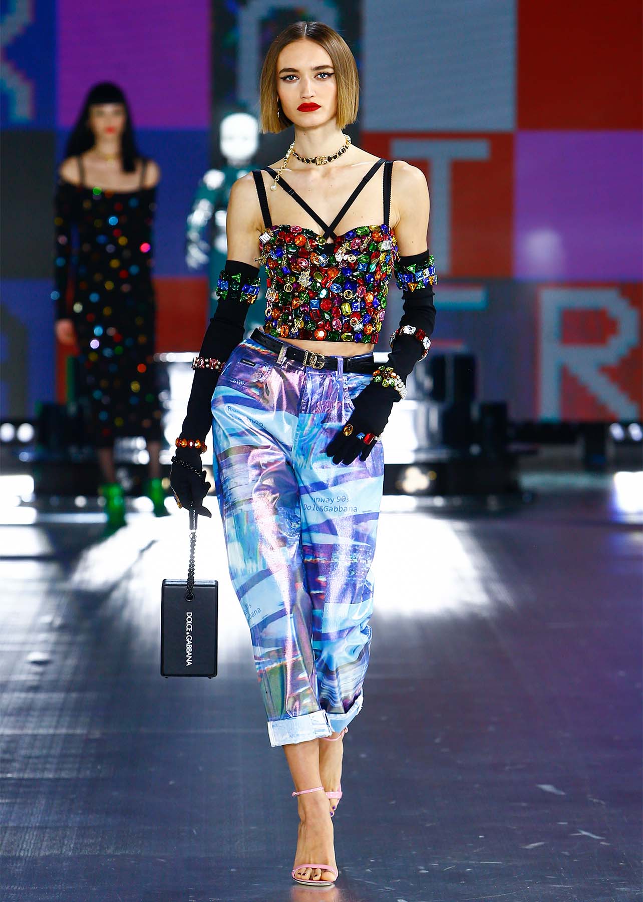 Woman walking on runway in a rainbow embellished crop top bustier, blue patterned trousers, heeled sandals and black gloves