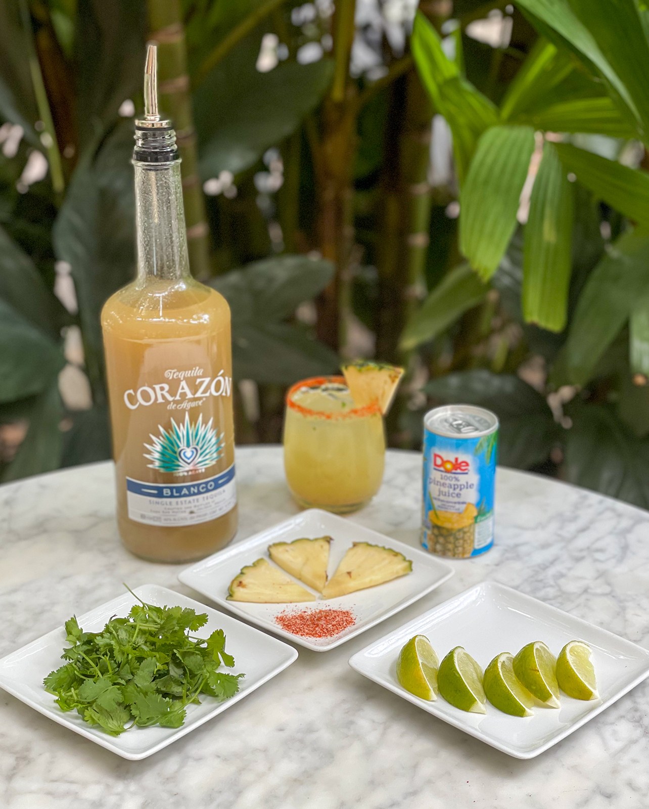 Image of the cocktail ingredients including cilantro, pineapple, limes, tahin, pineapple juice and a bottle of tequila