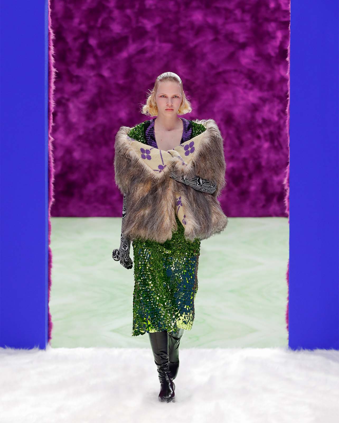 Woman walking a runway in a fur shawl, green sequin dress and black knee-high boots