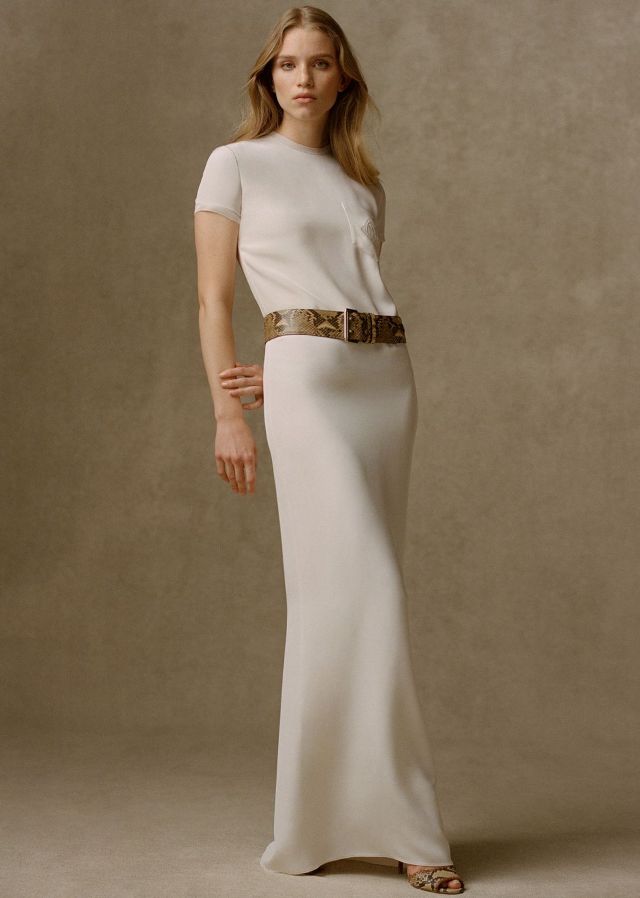 Woman photographed in an ivory floor length dress paired with a snakeskin waist belt and matching sandals