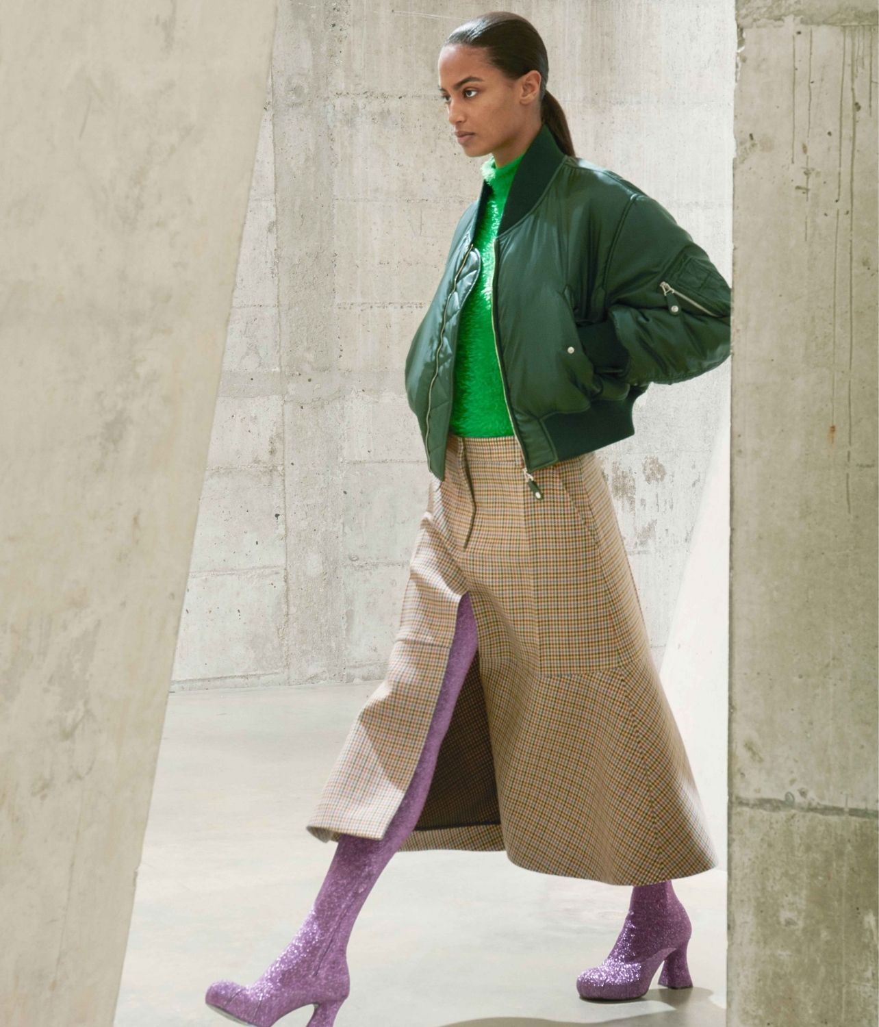 Woman walking on runway in Stella McCartney with green shirt and jacket, paired with a plaid skirt and thigh high purple glitter boots