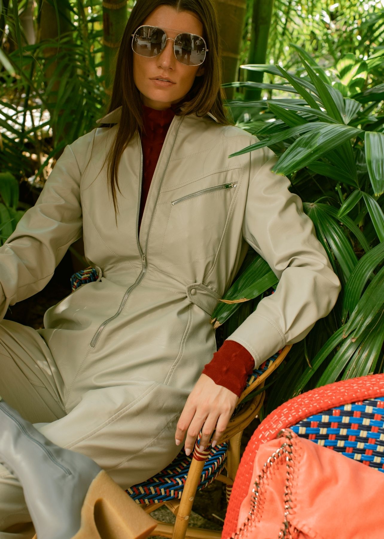 Woman sitting on a chair in a tan faux-leather jumpsuit with red turtleneck, grey boots and aviator sunglasses