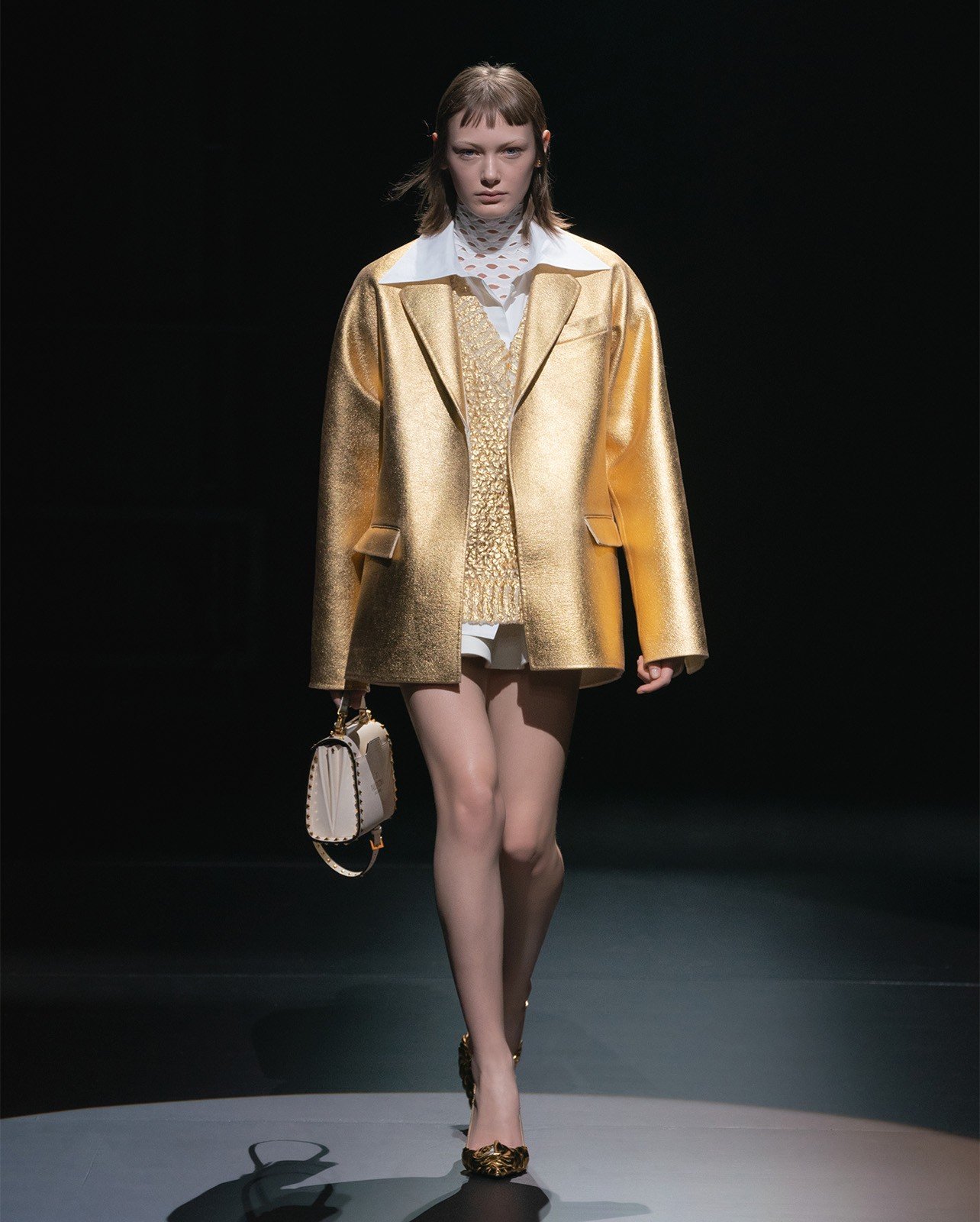 Woman walking on a runway wearing a gold jacket over a gold sweater and white undergarments