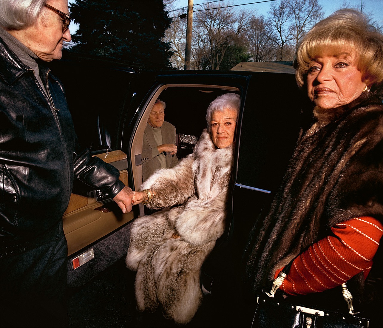 Photograph of an elderly man helping an elderly woman out of a car, dressed in an opulent fur coat