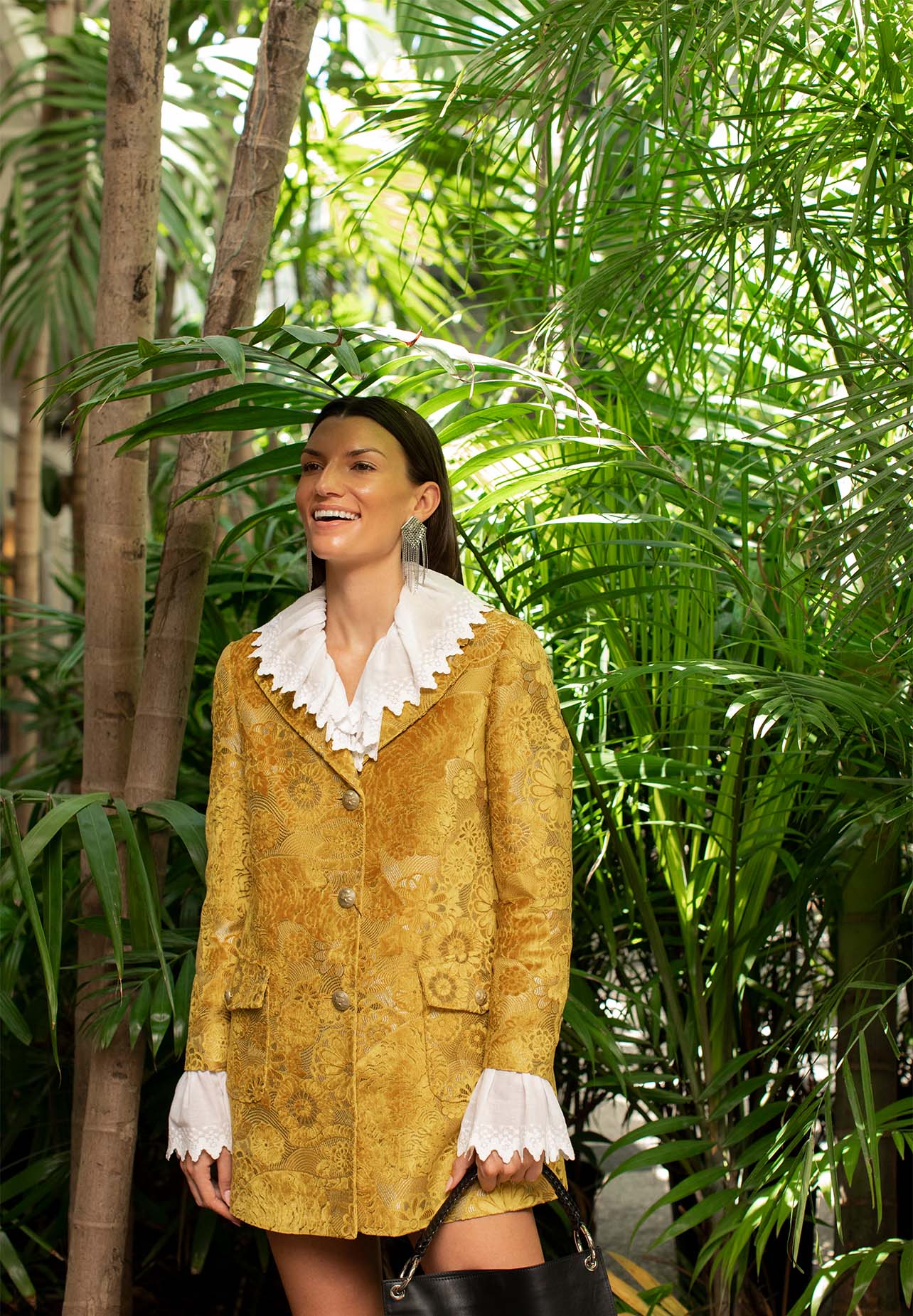Woman smiling in front of palm trees wearing a yellow Etro coat with a white blouse underneath