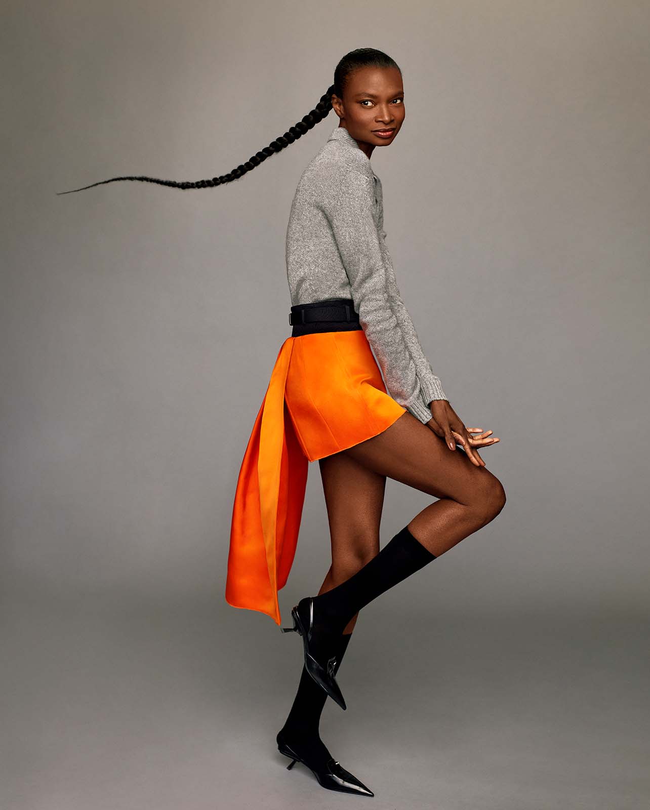 Model poses in a grey sweater and orange skirt, black high socks and heels