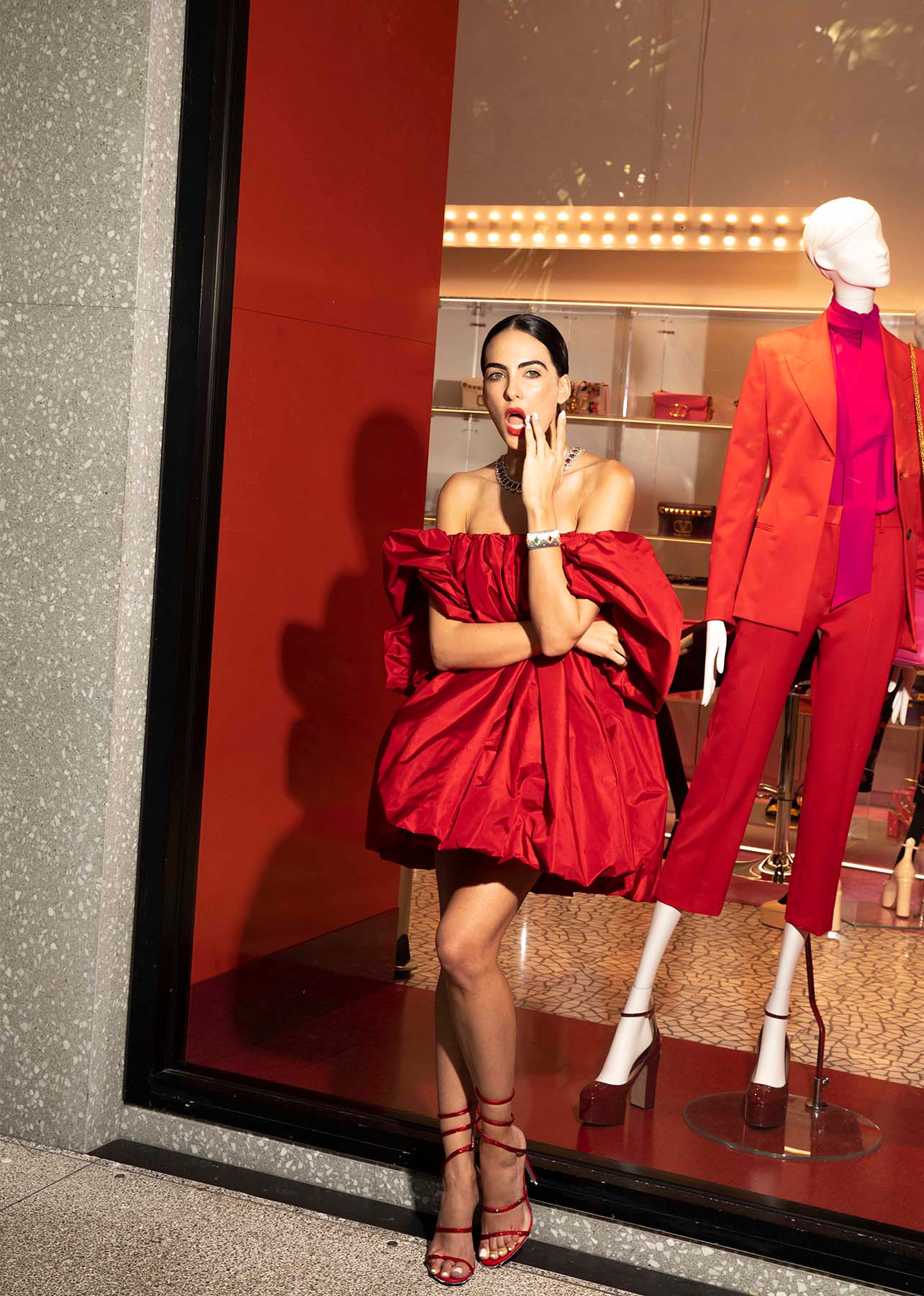 Model leans against the Valentino storefront in a red Valentino mini dress