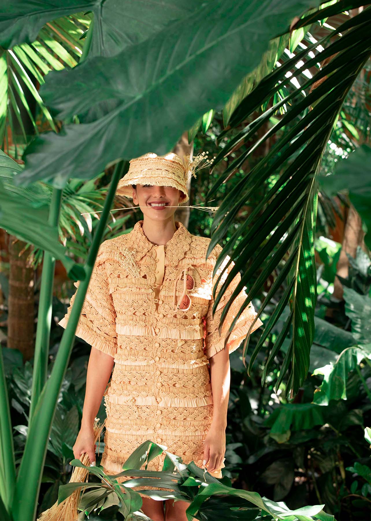 model stands among the palm trees in a full raffia look, holding a piece of straw in her mouth