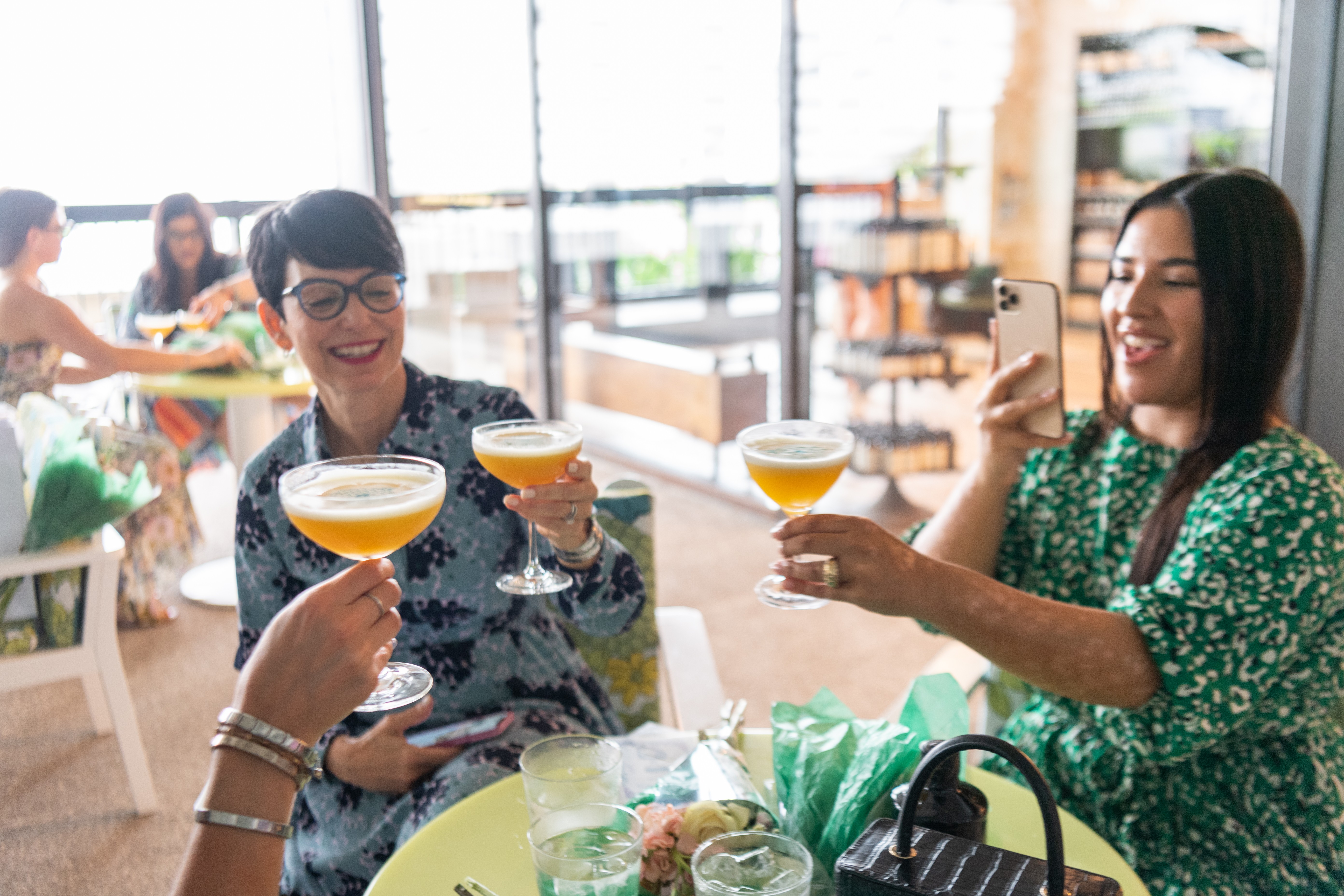 Three women cheers their cocktails while one woman takes a photo
