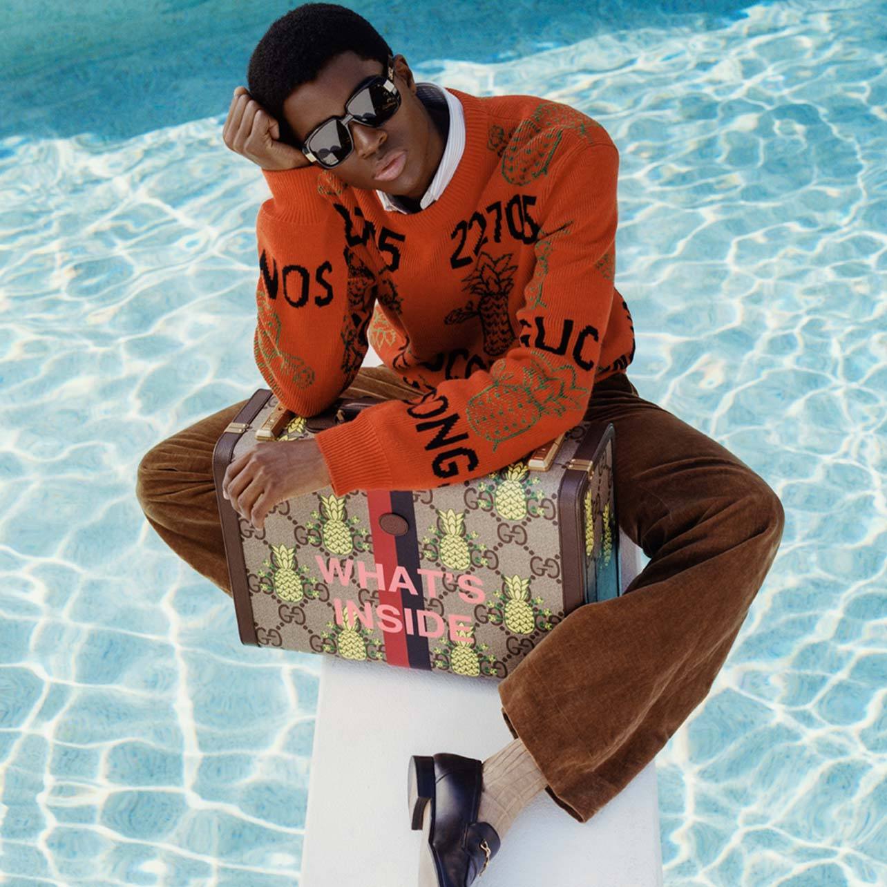 Man sitting on a diving board over a pool holding a Gucci monogrammed suitcase