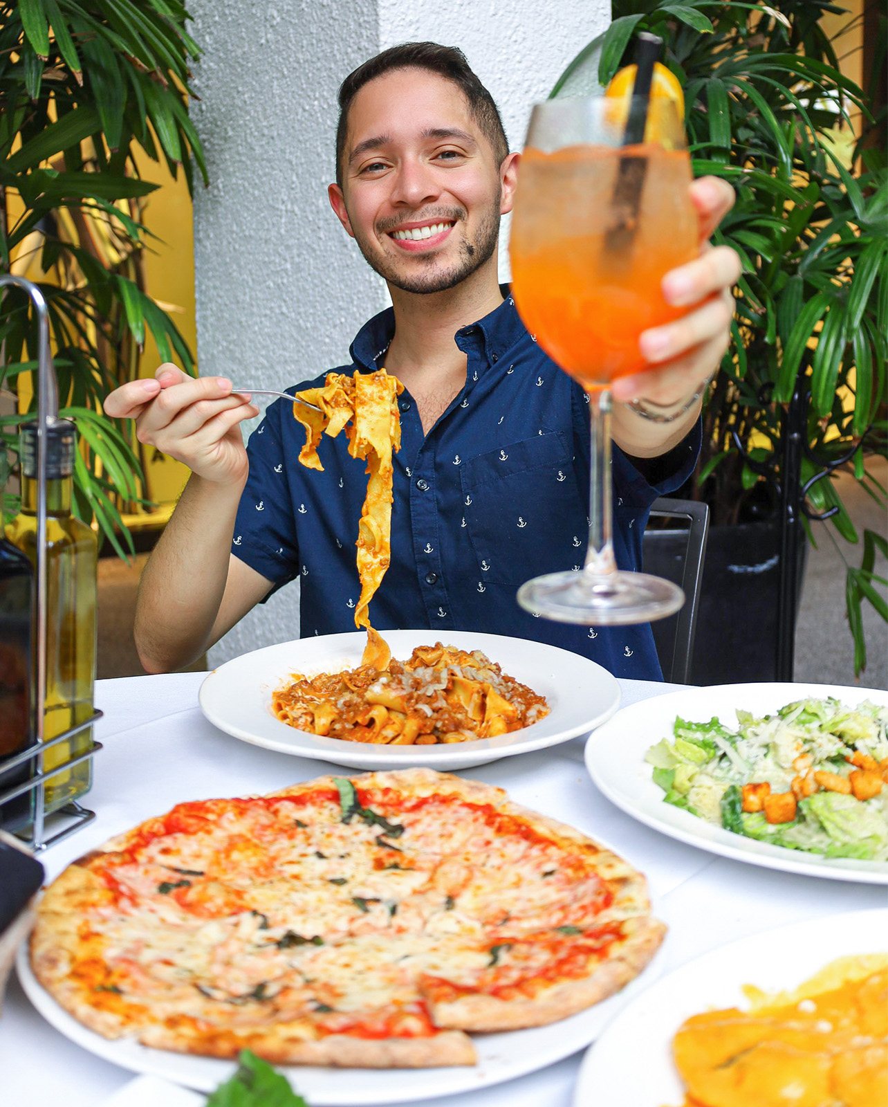 Man smiles with a cocktail and a dishes of pizza and pasta in front of him