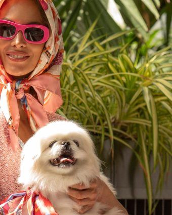 Model smiles with dog wearing sunglasses and a scarf wrapped around her head