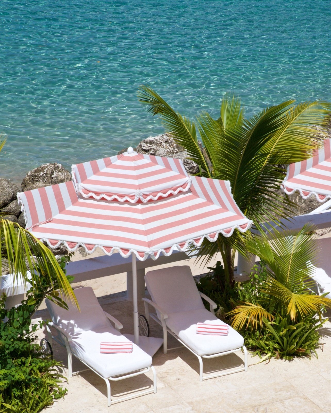 Two beach chair perched abover the water with a pink and white striped umbrella and matching towels
