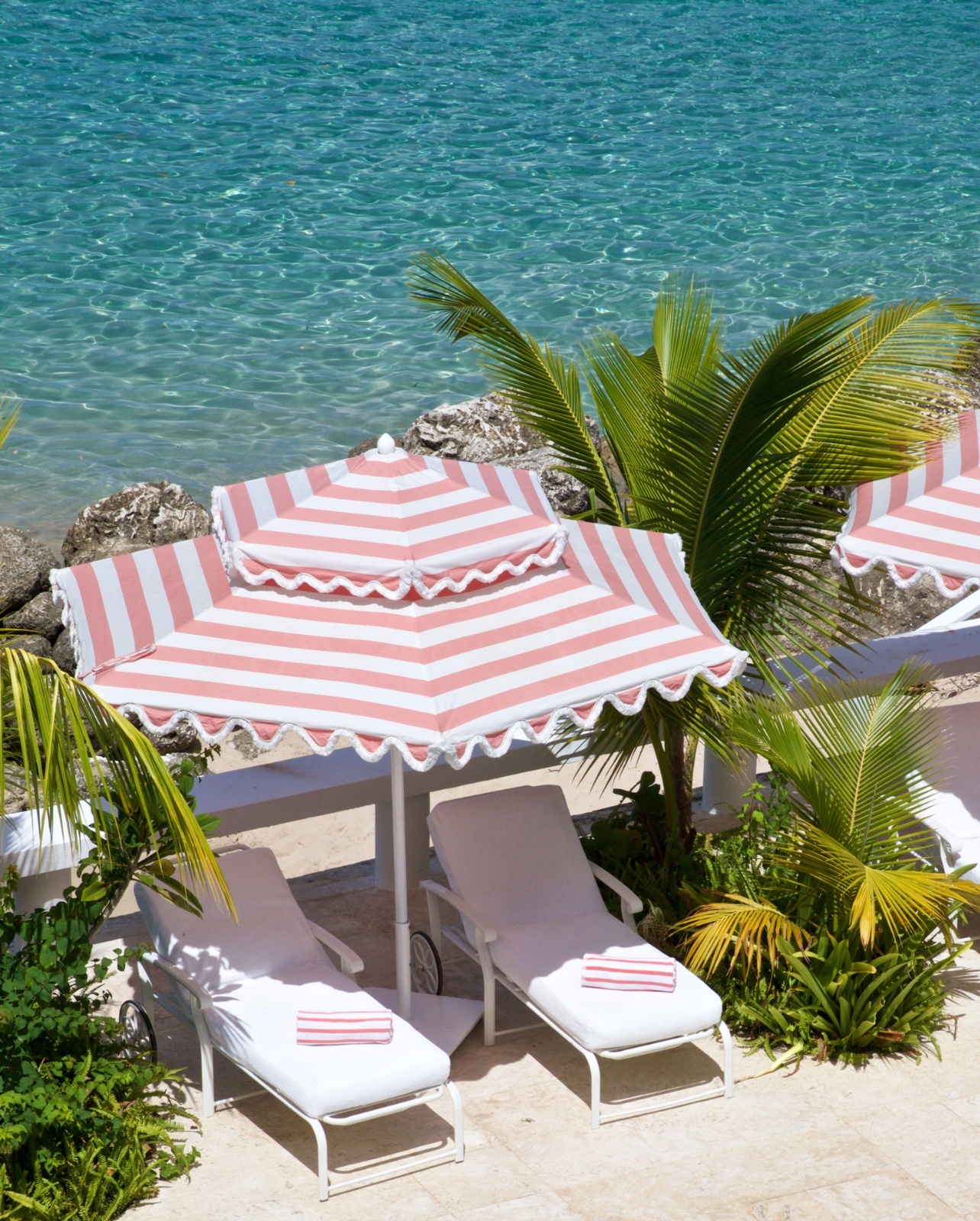 Two beach chair perched abover the water with a pink and white striped umbrella and matching towels