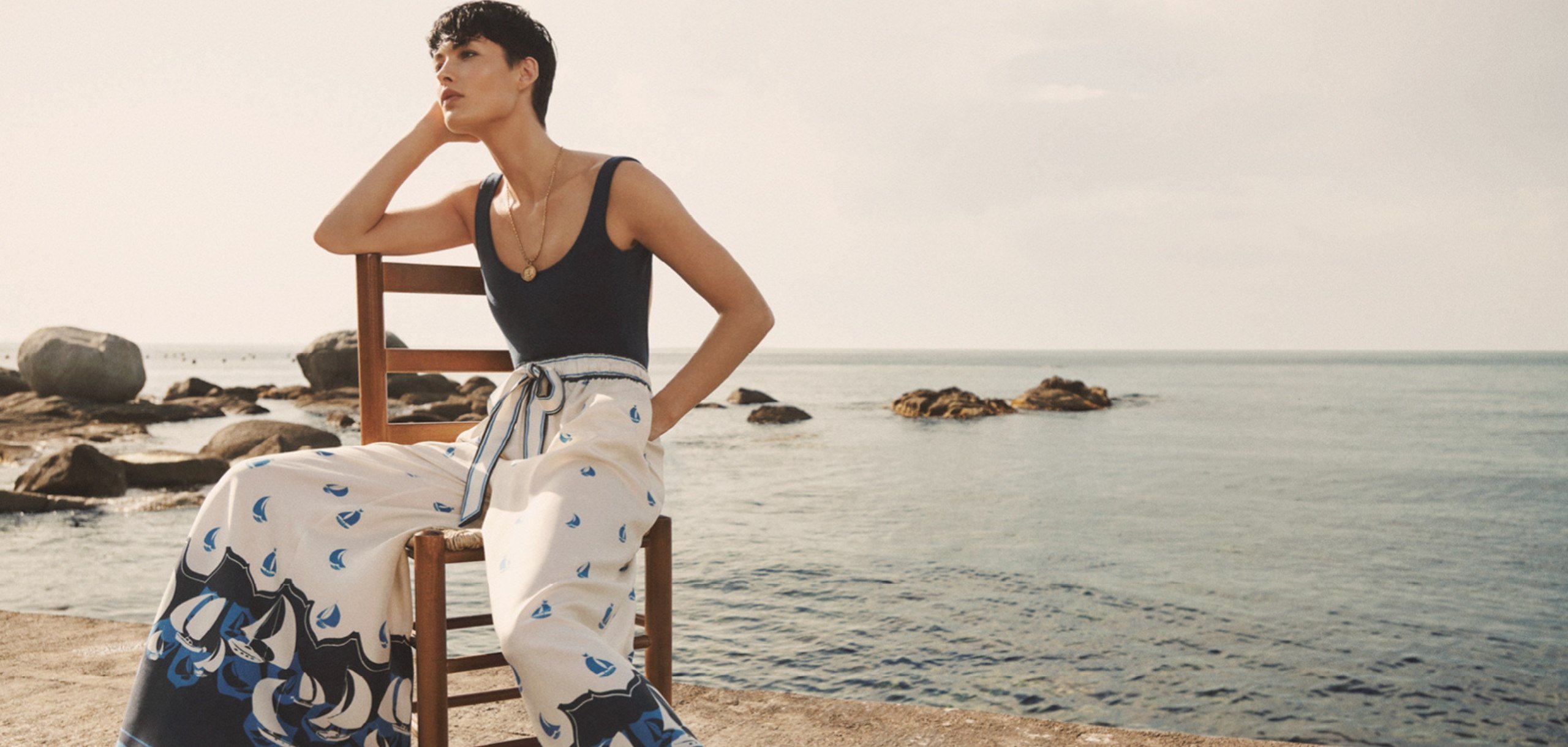Model sitting on a wood chair at the beach dressed in a navy sleeveless top and sailboat print trousers