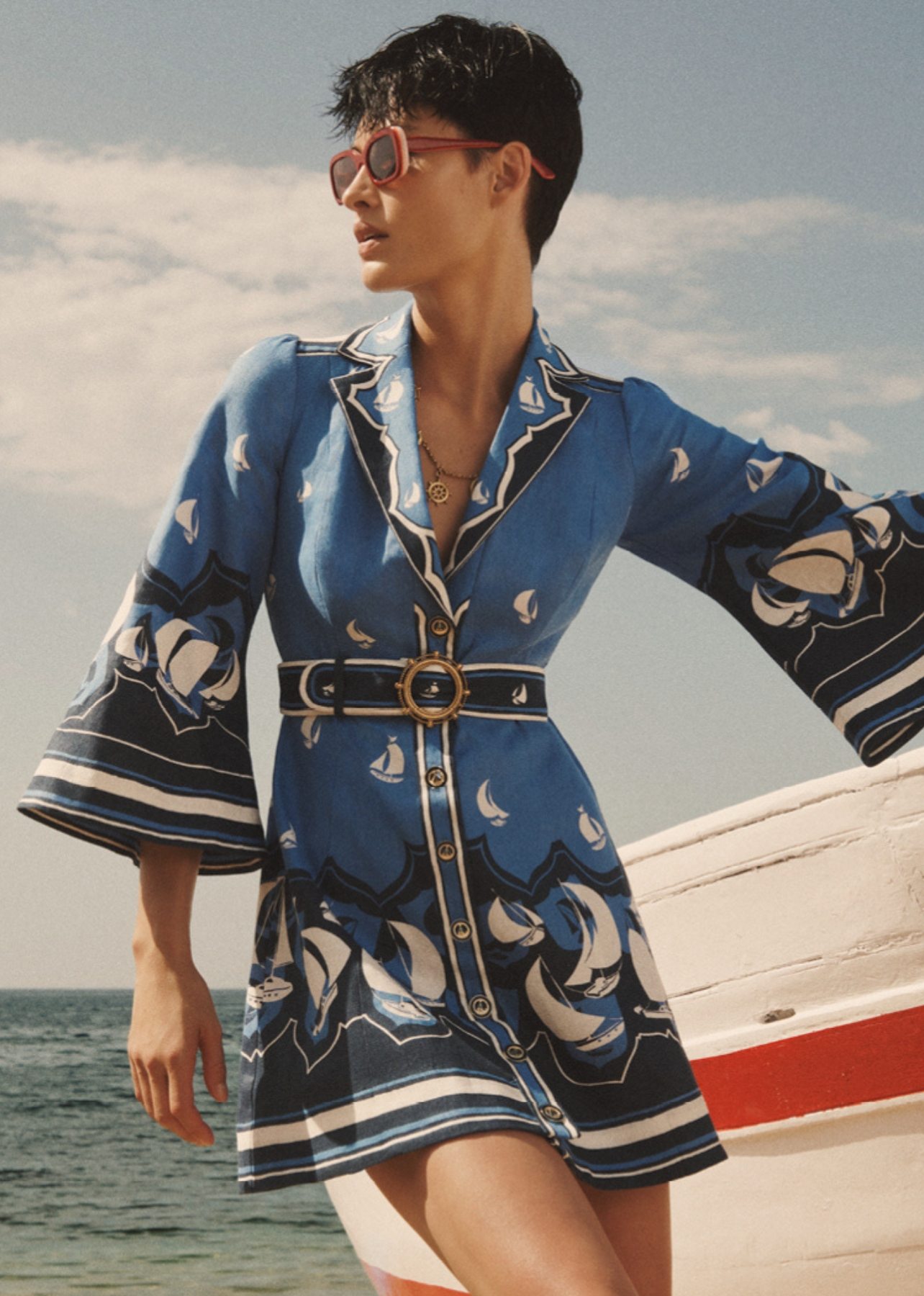 Model posing on the beach dressed in a blue sailboat print mini dress with belt detailing