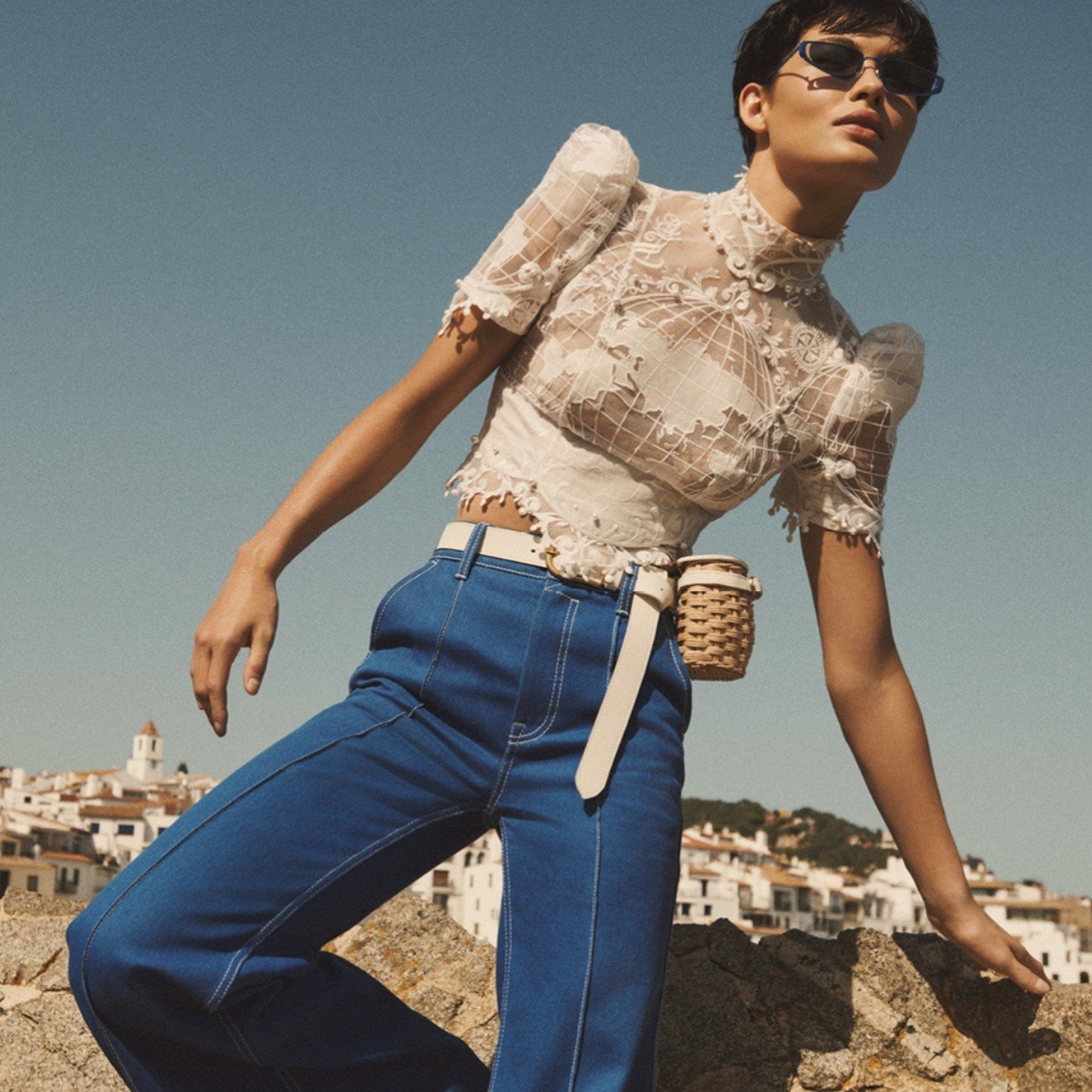 Model dressed in an embroidered blouse with map detailing, denim pants, and a woven basket belt bag