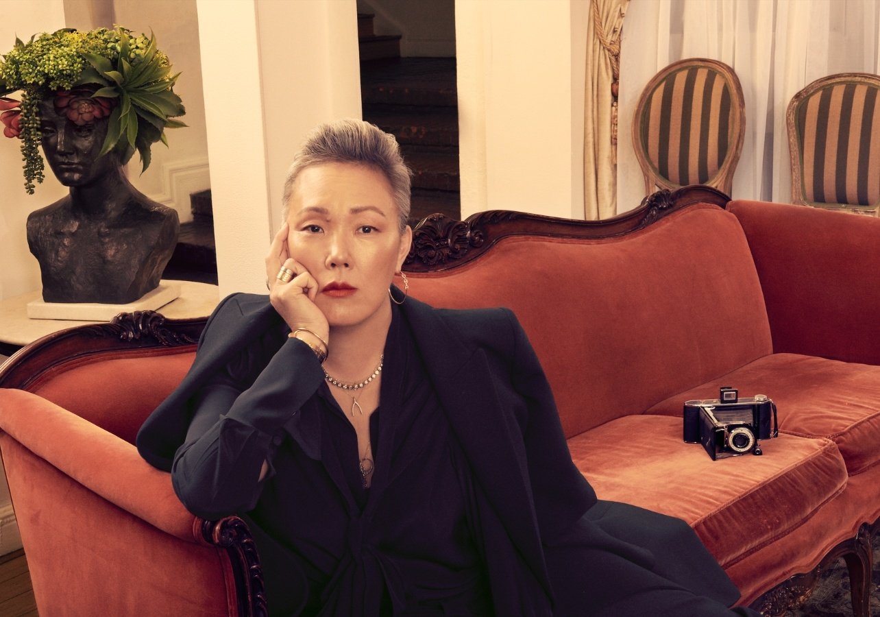 Jeanne Yang sitting on couch with a camera positioned beside her. Styled in a black button down and draped black blazer with gold jewelry