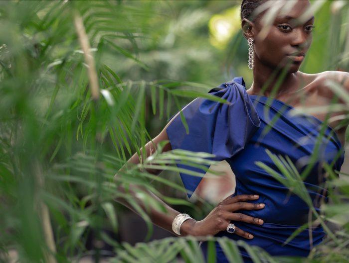 Model behind tropical foliage dressed in a royal blue one shoulder dress and diamond and sapphire jewelry.