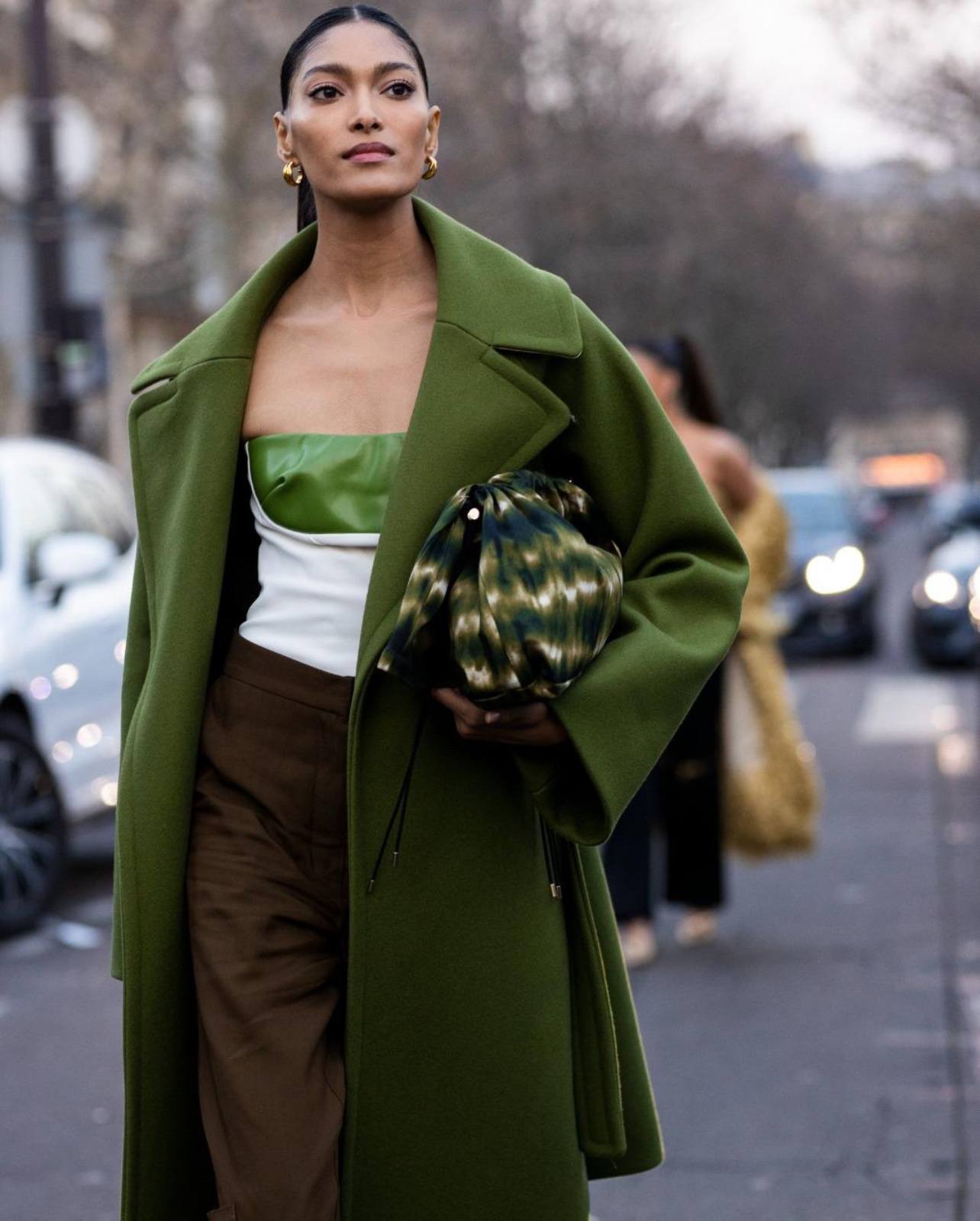 Model Pritika walking wearing a green wool coat, brown trousers, and a large handbag