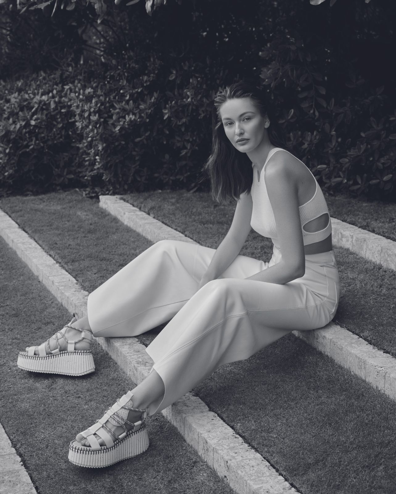 Model poses in grass wearing white Chloe pants, top, and platform sandals