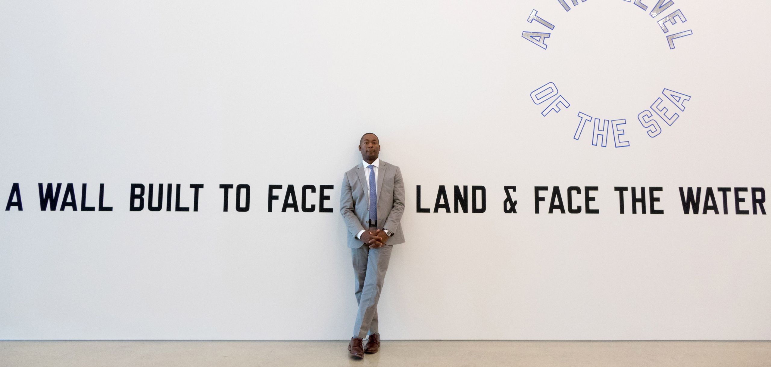 Portrait of Franklin Sirmans wearing a grey suit in front of Laurence Weiners A Wall Built to Face the Land & Face the Water at the Level of the Sea