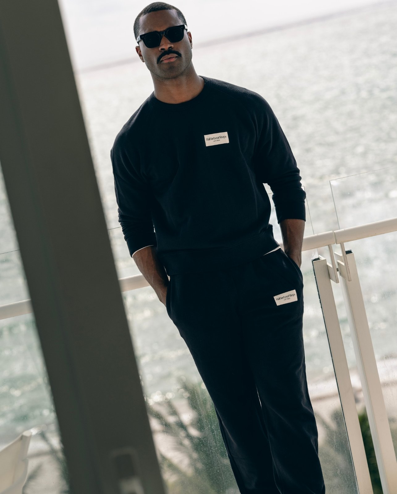 Lifestyle shot of male model styled in the black Bal Harbour Shops sweatsuit on balcony overlooking the ocean
