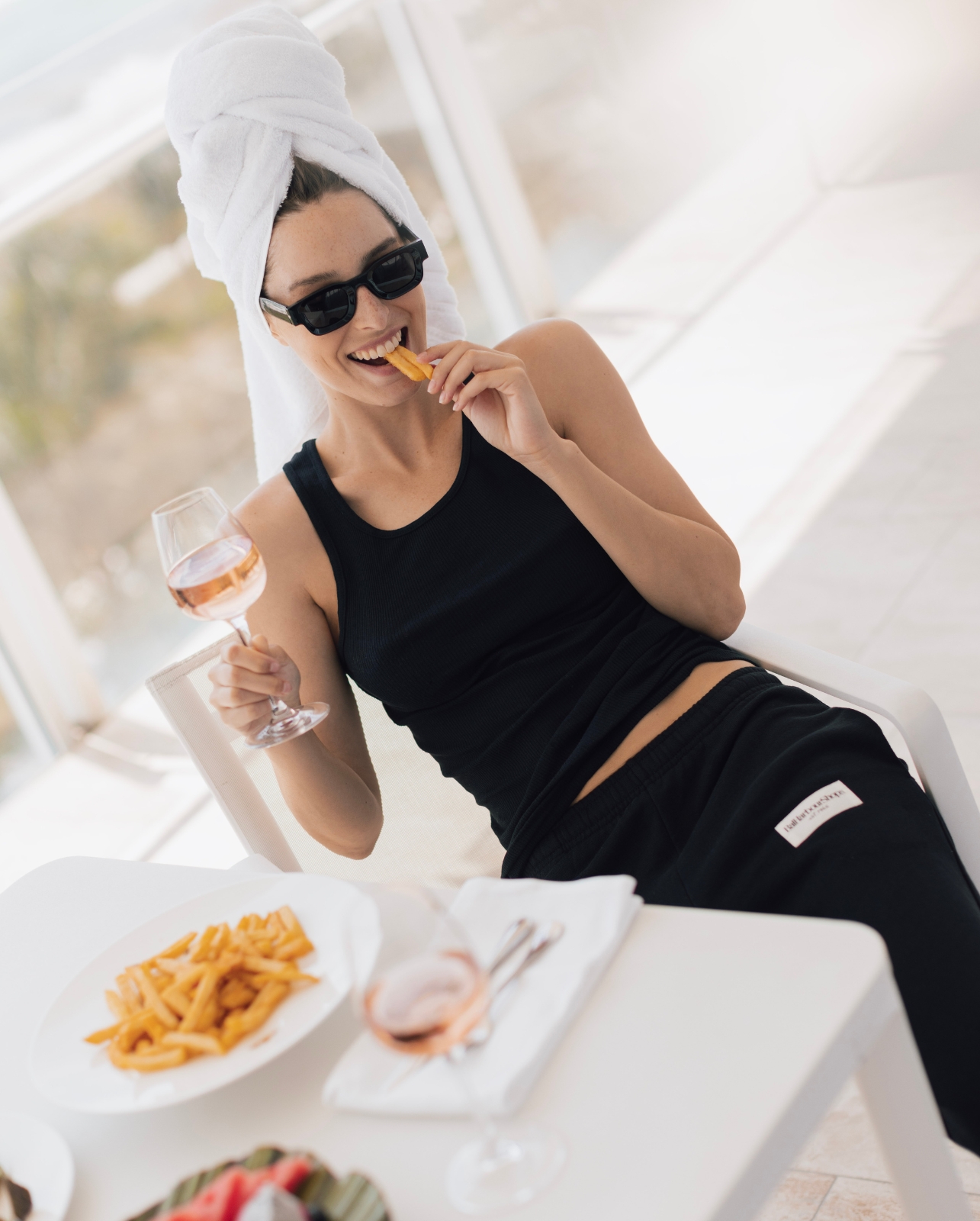 Lifestyle shot of female model styled in the black Bal Harbour Shops 1965 sweatpants, dining and sipping rose on a balcony over-looking the ocean