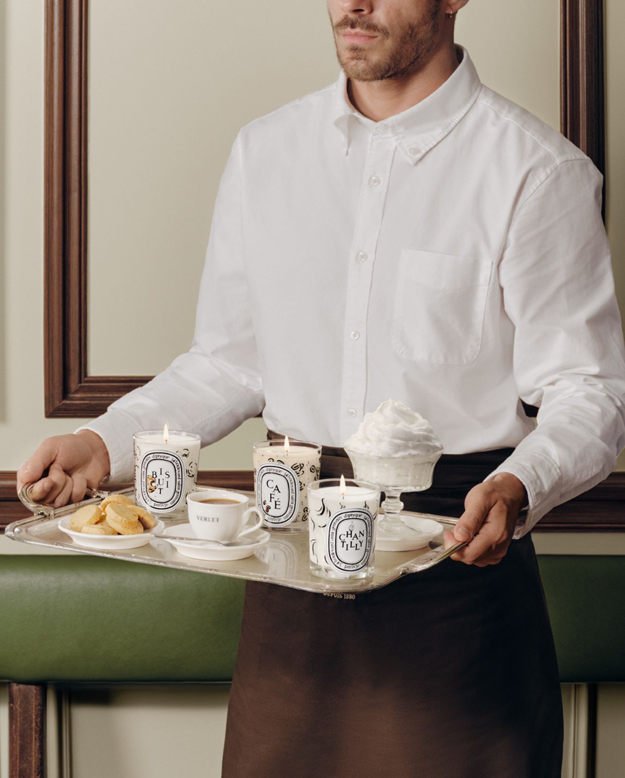 Man carrying a tray of Diptyque candles from the Café Verlet collection along with coffee and cookies