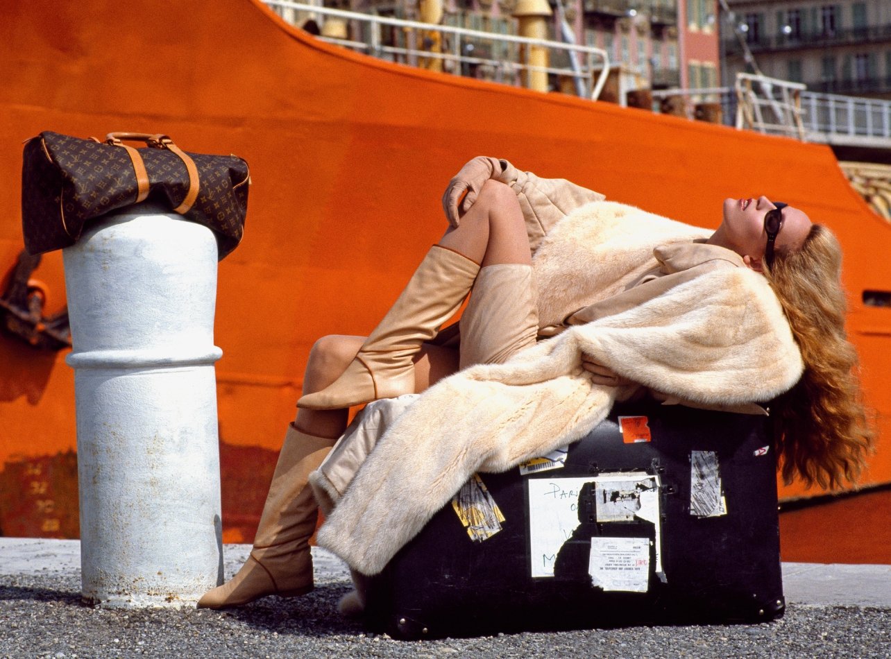 Model with Louis Vuitton travel duffel wearing a fur coat, leather boots and sunglasses posing in front of an orange boat