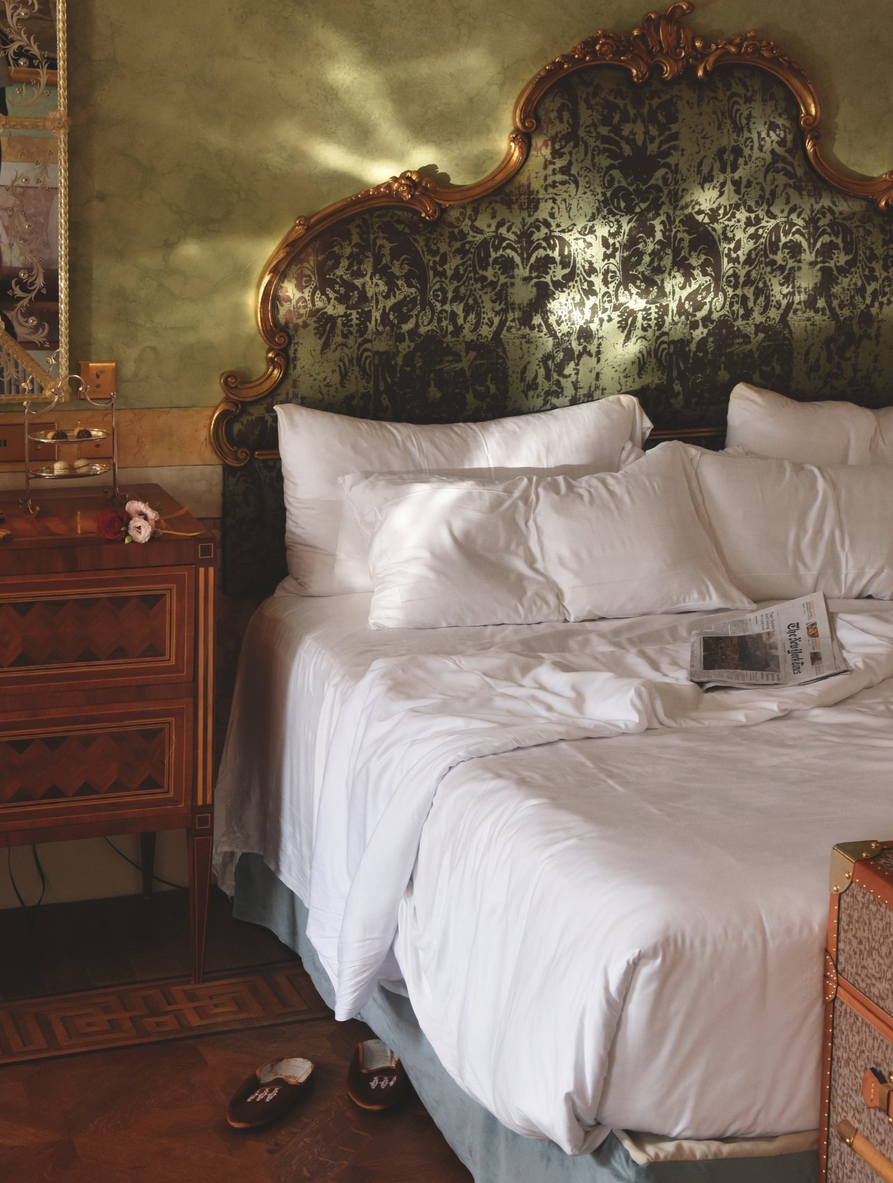 A bedroom at the Passalacqua hotel featuring wood furniture, gold details, green wall accents, and a newspaper resting on a bed with white sheets.