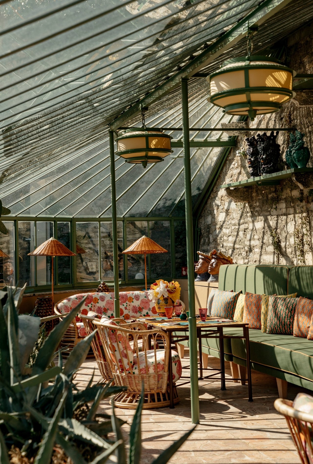 A sunlit interior of the Passalacqua hotel’s window-filled sunroom featuring a green couch, floral rattan chairs, and a dining table set for guests