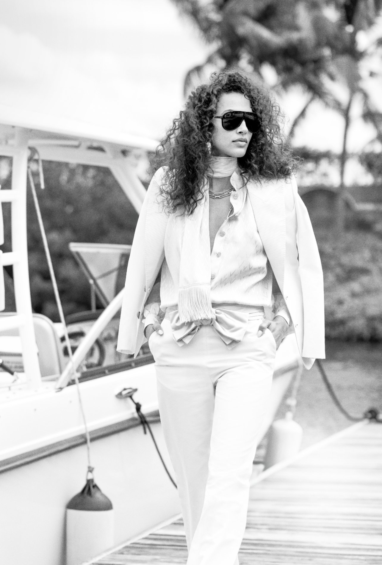 Fashion model in an all-white Valentino outfit—blazer, scarf, shirt, and pants—posing beside a boat, wearing Tom Ford sunglasses