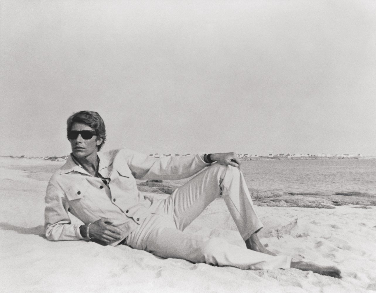 Male model wearing a white jacket, matching pants, and sunglasses poses on a beach, from the book Yves Saint Laurent and Photography