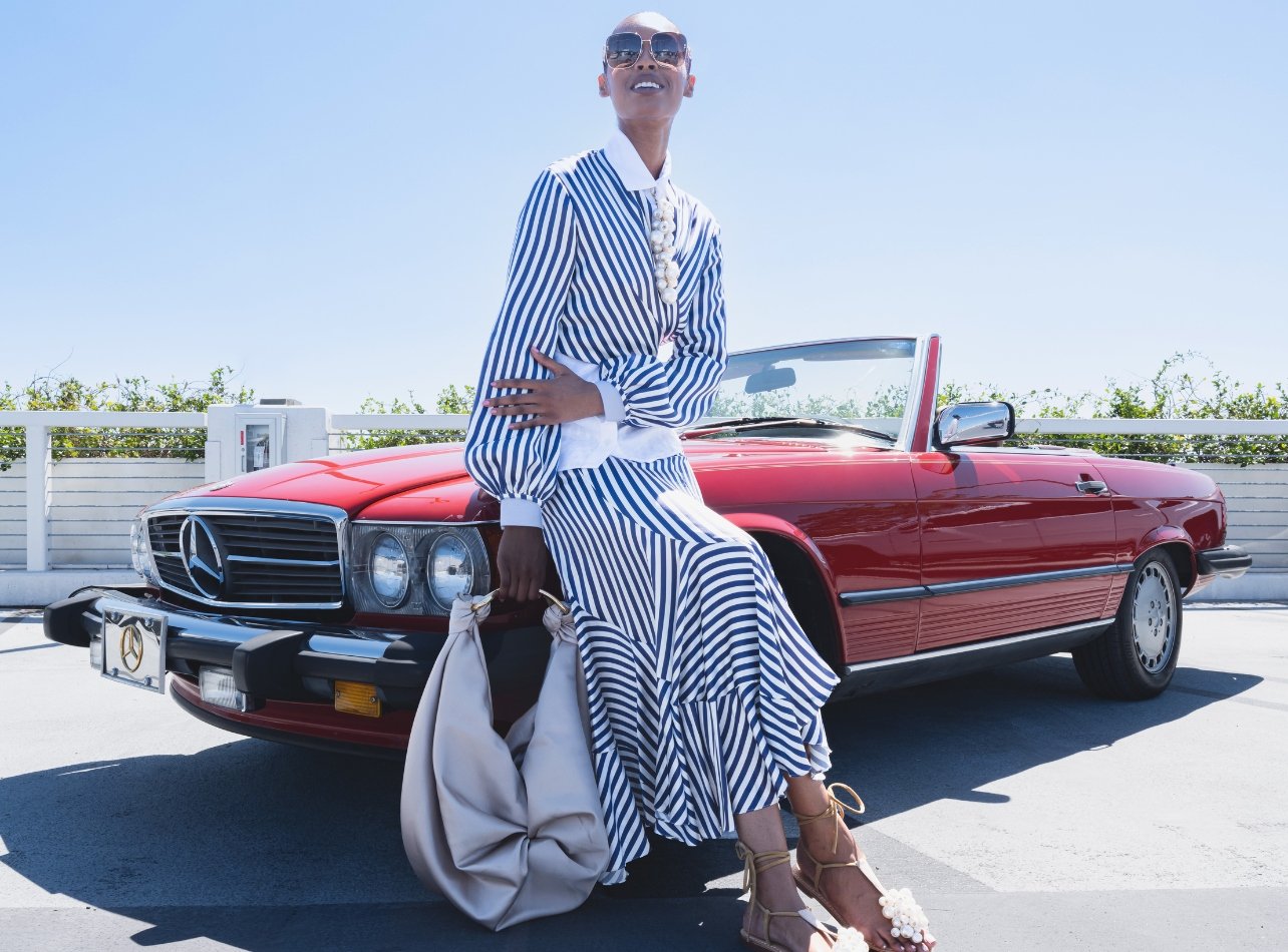 A model posed in front of a red convertible wearing a blue and white striped dress holding an oversized silver bag