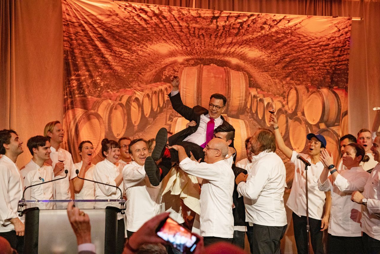 A man wearing a suit and tie is tossed in the air surrounded by chefs in white uniforms