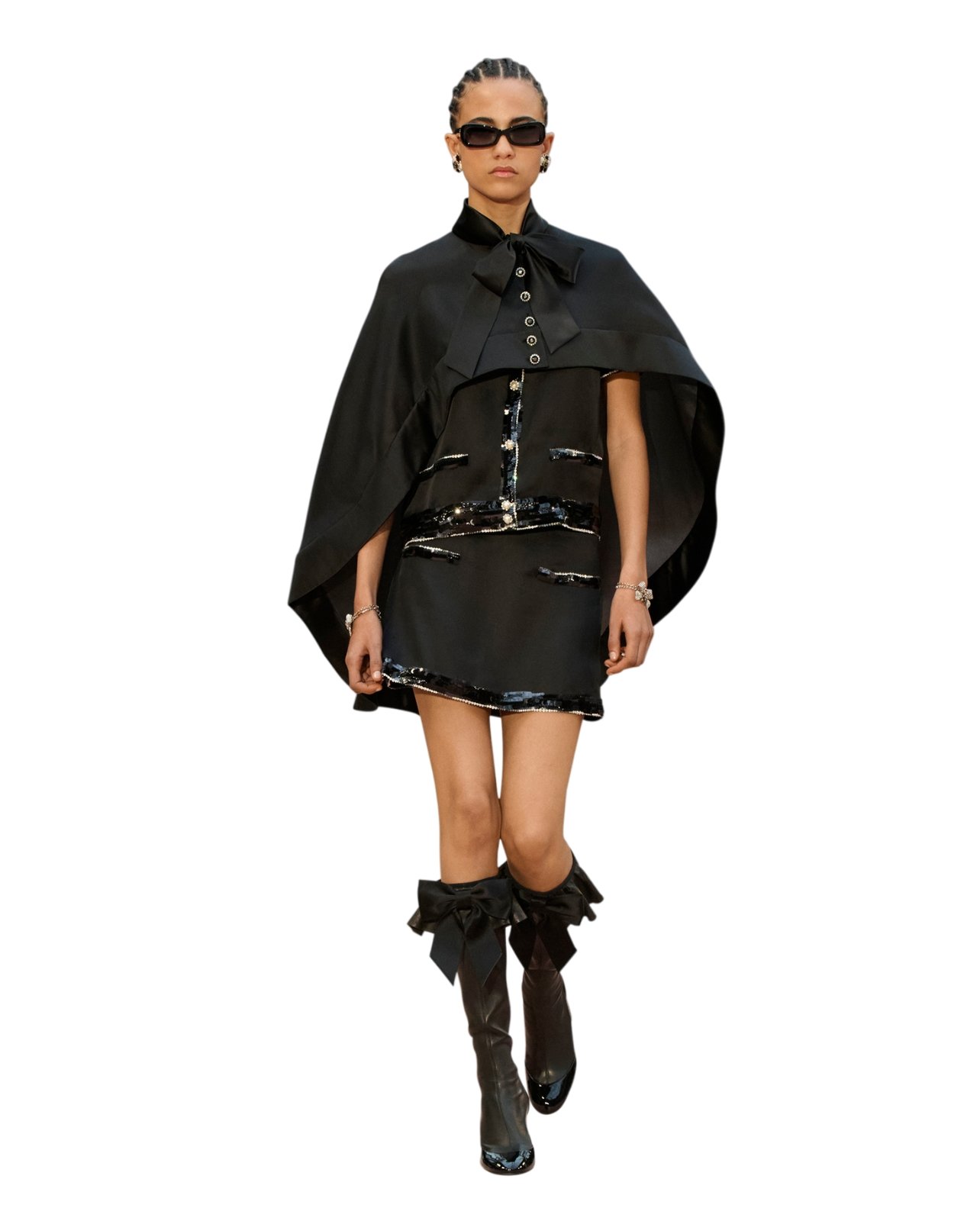 Runway model wearing a black dress, cape, tall boots and sunglasses