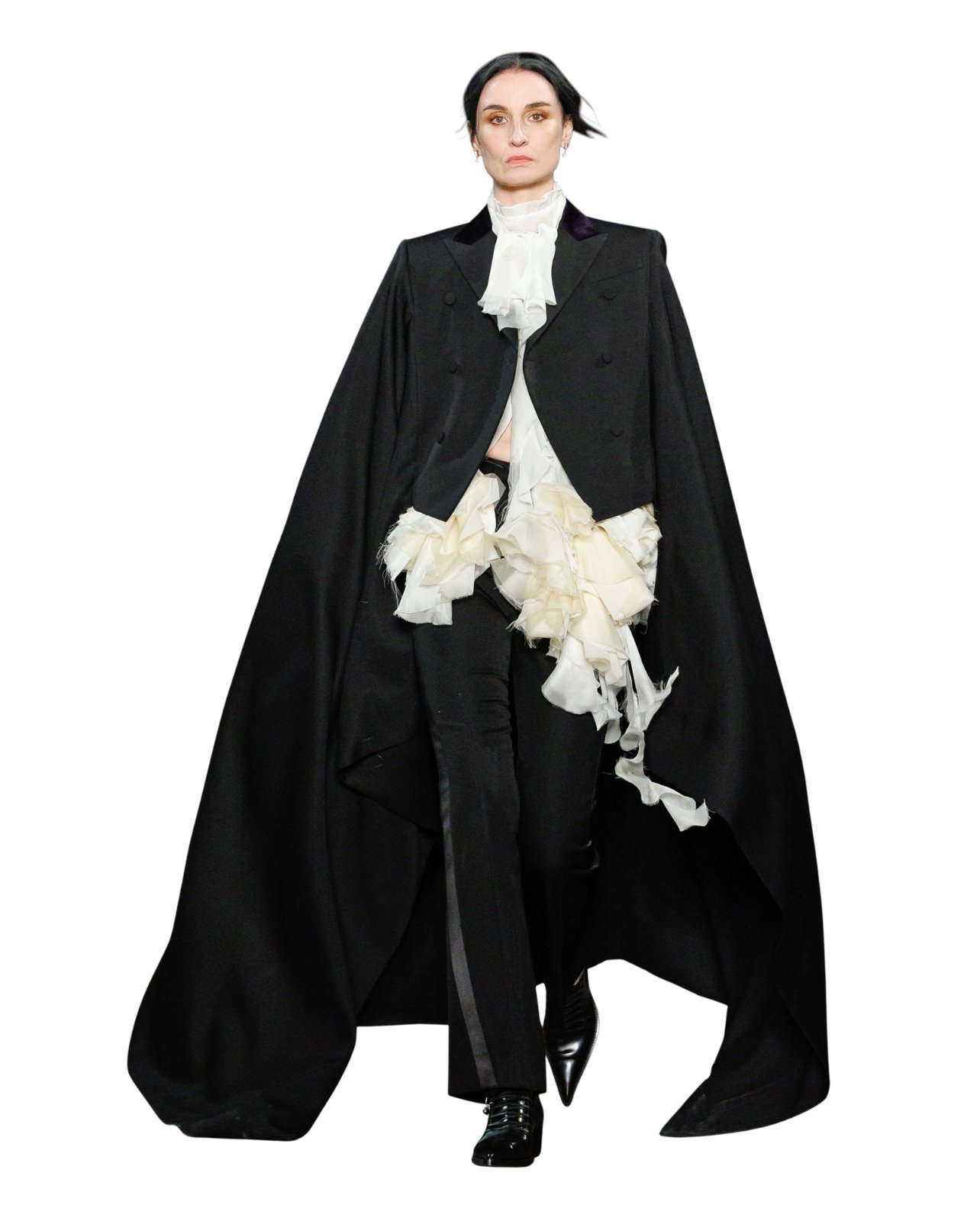 Runway model wearing a floor length black cape with white ruffles and black pants