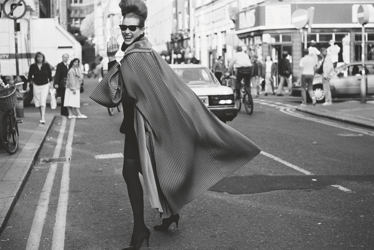 A black and white photo of a fashion model walking in the street wearing a long fan coat, sunglasses, and black heels