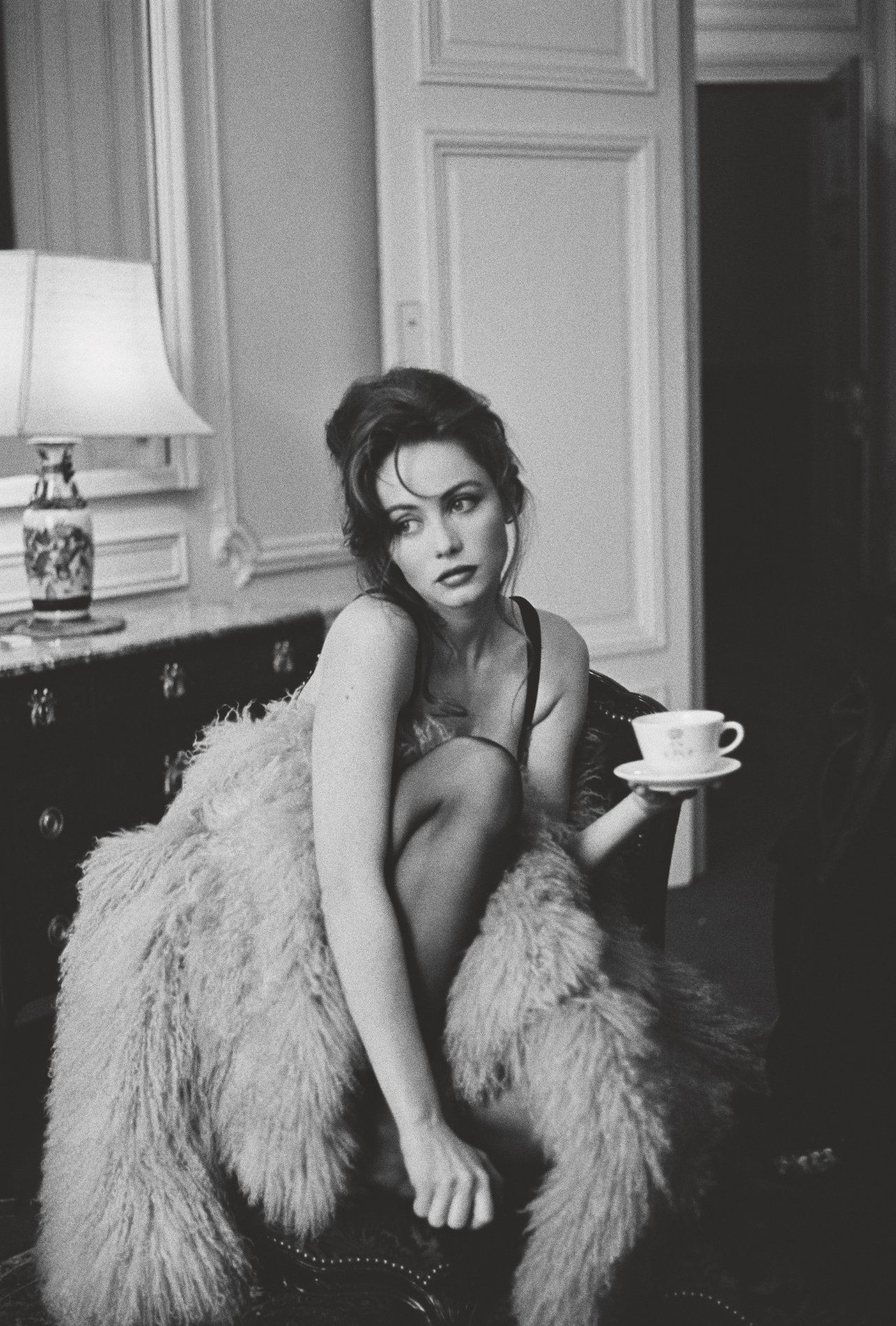 A black and white photo of a fashion model draped in fur on a couch with a coffee cup