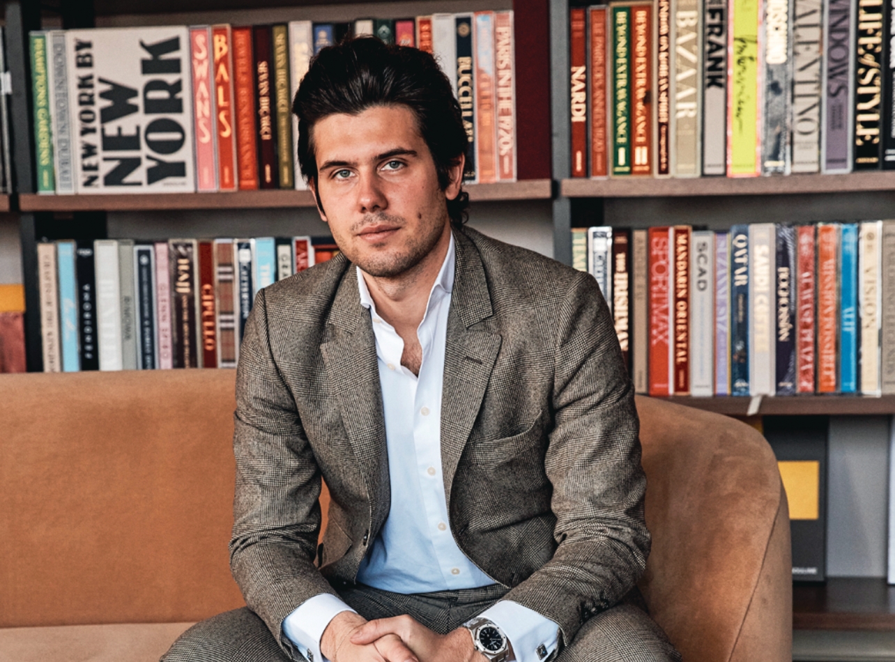 A portrait of Alexandre Assouline sitting on a couch with a bookcase behind him