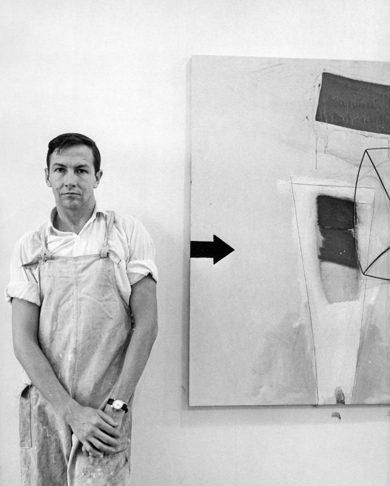 A black and white portrait of Robert Rauschenberg standing next to a painting in his studio
