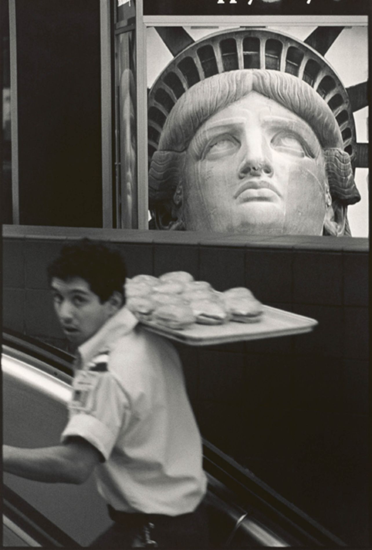 A black and white picture of a man walking upstairs carrying a tray of burgers with a statue of liberty artwork in the background